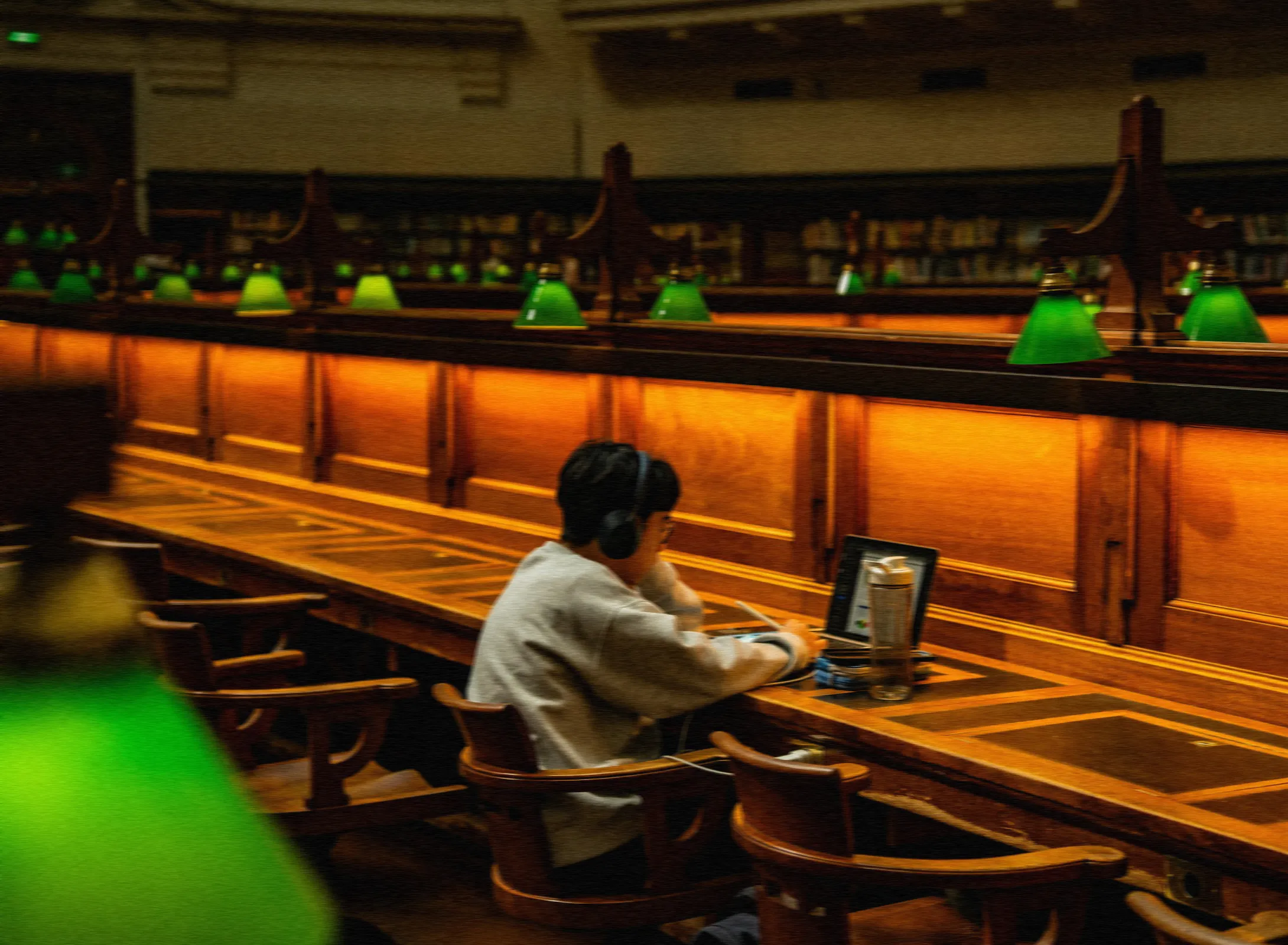 Um estudante de fone de ouvido estuda no notebook numa biblioteca, sob luminárias verdes